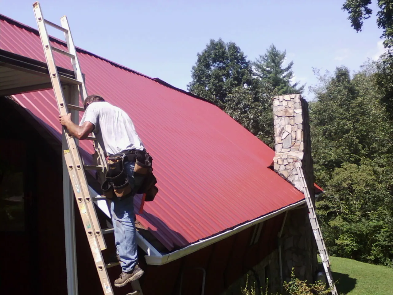 Roofer on a ladder beside a red metal roof and stone chimney — Soffit & Fascia Repair by Roofing Brothers in Kingsburg