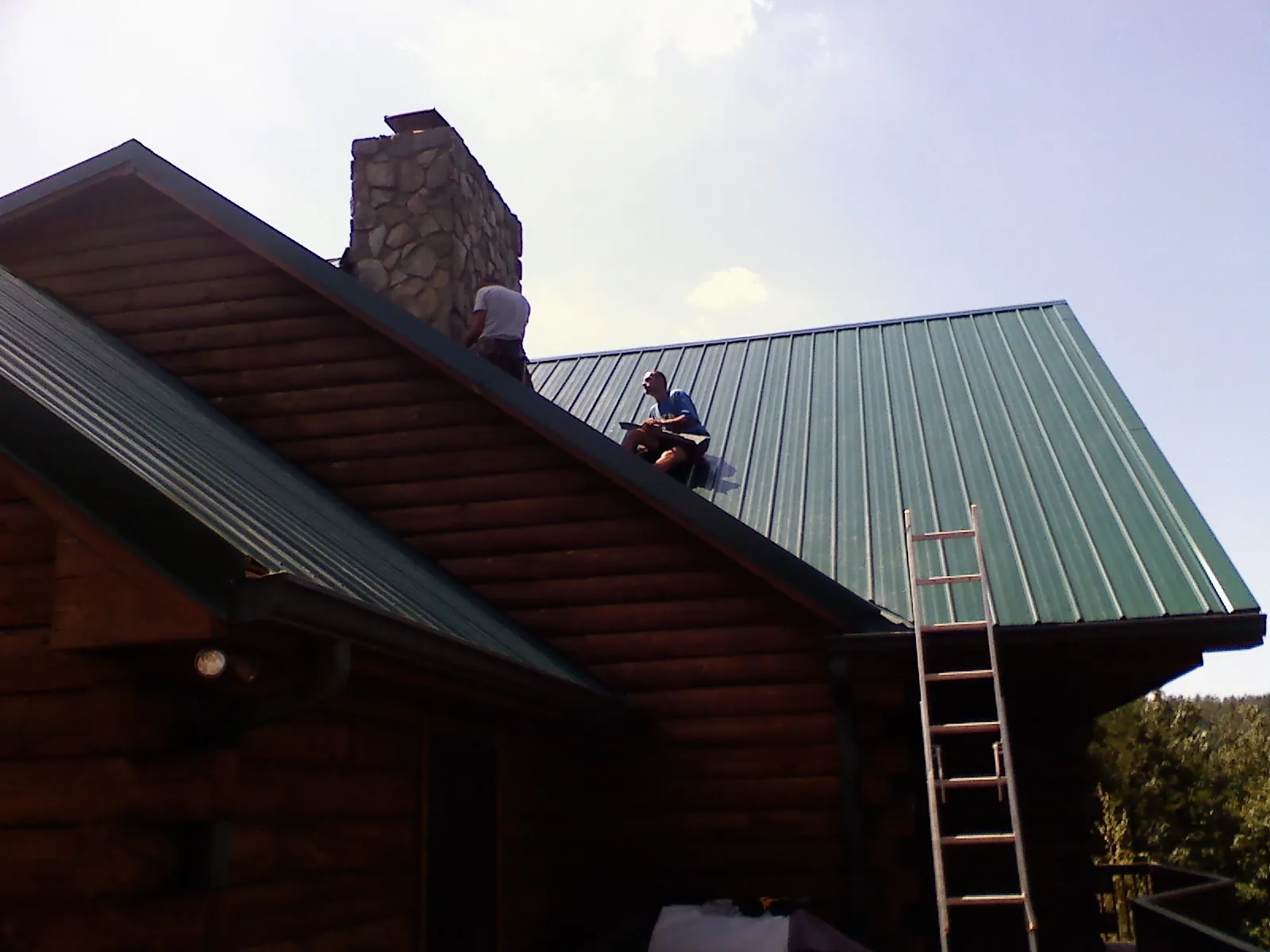 Roofers installing green metal panels beside a stone chimney on a log home in Kingsburg